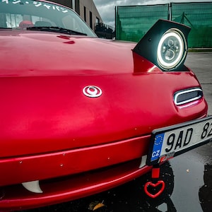 以下が含まれることがあります： 白いナンバープレートに「9AD 82 CZ」と表示された赤いマツダ・ロードスター。車には白いハート型の牽引フックと、フロントガラスに「サン・ジョイ」と書かれた白いステッカーが貼られています。