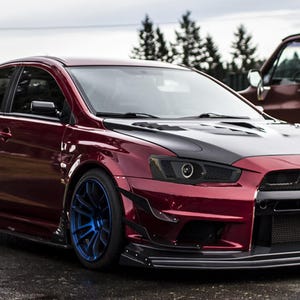 Op de afbeelding: Een aangepaste bordeauxrode sedan met een zwarte motorkap en blauwe velgen. De auto heeft een grote achtervleugel en een aangepaste carrosseriekit. Een bordeauxrode pick-up truck staat op de achtergrond.