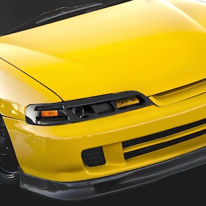 May include: A bright yellow car front end featuring a sleek bonnet, black mesh grille inserts, and integrated amber turn signal lights. The vehicle includes a dark carbon fibre style front lip spoiler and black wheels, showcasing a streamlined design.