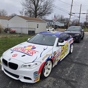 May include: A white BMW saloon with a Wonder Bread racing livery. The car has a yellow number 25 on the side and a blue and red flame design. The car is parked on a driveway with a black Jeep Cherokee behind it.
