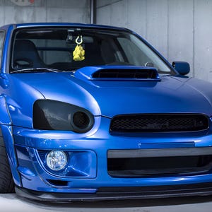 May include: A blue Subaru Impreza WRX with blacked-out headlights and a black mesh grille. The car has a black front lip spoiler and a yellow keychain hanging from the rearview mirror.