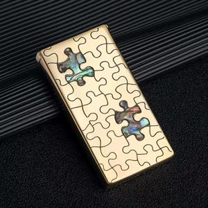 May include: A gold-coloured lighter with a puzzle design. The lighter has a series of puzzle piece outlines, with two pieces filled with iridescent abalone shell. The lighter is rectangular and has a smooth, reflective surface.