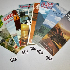 May include: A collection of vintage Alberta road maps, fanned out in a rainbow pattern, with the years 50s, 60s, 70s, 90s, and 00s written on white labels below.