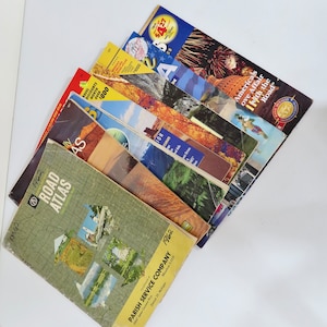May include: A stack of vintage road atlases and travel guides, including a Parish Service Company Road Atlas, a 1962 Road Atlas, and a guide titled "America's Love Affair with the Road".