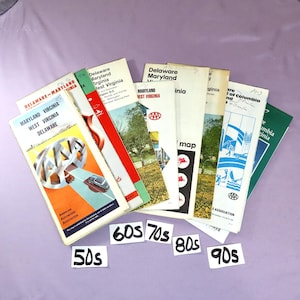 May include: A stack of vintage AAA road maps from the 1950s through the 1990s. The maps are in various shades of blue, red, and green, and feature the AAA logo. The maps are labeled with the states they cover, including Delaware, Maryland, Virginia, and West Virginia.