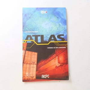 May include: A red, white, and blue Canadian Railway Atlas book with the title "ATLAS" in yellow letters. The cover features a train speeding through a tunnel with cargo containers labeled "53ft" and the text "CHEMINS DE FER CANADIENS".