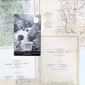 May include: A collection of vintage maps and brochures, including a black and white image of a road with cars and the text "CACHE NATIONAL FOREST". Other documents include "FISHLAKE NATIONAL FOREST" and detailed maps.