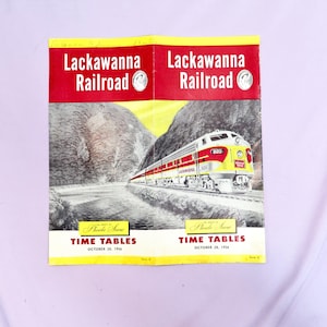 May include: A vintage Lackawanna Railroad time table booklet with a yellow and red cover. The cover features a photo of a train traveling through a mountain valley. The text on the cover reads "Lackawanna Railroad" and "Phoebe Snow Time Tables, October 28, 1956."