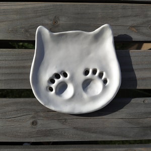 Op de afbeelding: Witte keramische schotel in de vorm van een kat met twee in de oppervlakte gegoten potafdrukken.