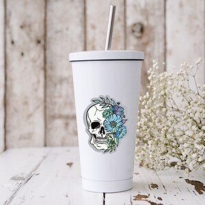 May include: White stainless steel tumbler with a black lid and a sticker of a white skull with blue and green flowers on the front.