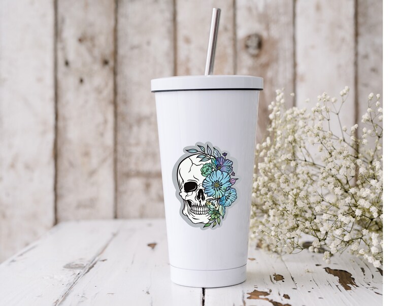 May include: White stainless steel tumbler with a black lid and a sticker of a white skull with blue and green flowers on the front.