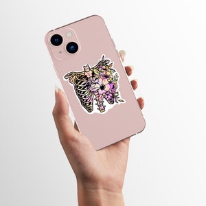 May include: A pink phone case featuring a sticker of a floral ribcage design. The sticker is in a watercolour style with pink, yellow and purple flowers.