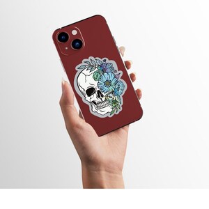 May include: A red phone case with a white skull sticker featuring blue and purple flowers. The skull is surrounded by leaves and vines.