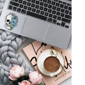 May include: A laptop computer with a sticker of a skull with flowers on it. The laptop is on a gray knitted blanket with pink flowers. A pink notebook with the text "GARANCE DOR&Eacute;" is in the background.