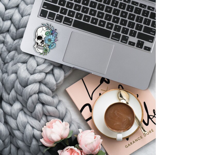 May include: A laptop computer with a sticker of a skull with flowers on it. The laptop is on a gray knitted blanket with pink flowers. A pink notebook with the text "GARANCE DOR&Eacute;" is in the background.