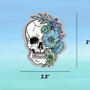 May include: A white skull with a floral design in shades of blue, green, and purple. The skull is 2.3 inches wide and 3 inches tall.