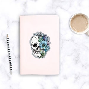 May include: A white skull with blue and green flowers covering one eye. The skull is outlined in black and the flowers are detailed with shading.