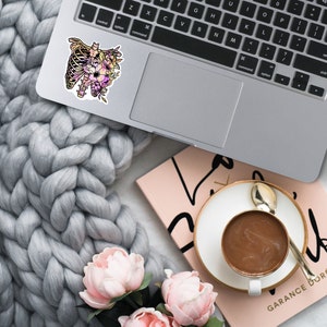 May include: A sticker featuring a floral design with a ribcage in the centre. The sticker is on a silver laptop computer. The laptop is on a grey knitted blanket. There is a pink notebook and a cup of coffee in the background.