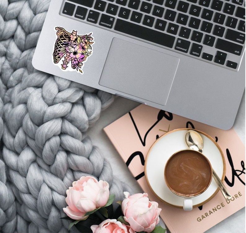 May include: A sticker featuring a floral design with a ribcage in the centre. The sticker is on a silver laptop computer. The laptop is on a grey knitted blanket. There is a pink notebook and a cup of coffee in the background.