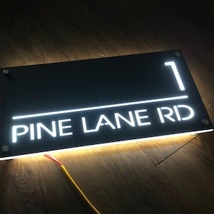 Pu&ograve; includere: Un cartello rettangolare nero con lettere LED bianche che recita "1 PINE LANE RD".