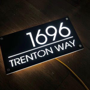 May include: Illuminated black address sign with the numbers "1696" and the text "Trenton Way" in white. The sign is rectangular and has a warm, glowing light emanating from behind the numbers and text.