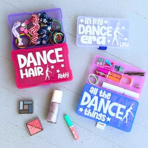 Puede incluir: Cuatro cajas de almacenamiento de plástico, dos abiertas, llenas de accesorios de baile. Una caja rosa dice "DANCE HAIR" con una silueta de bailarina y "Abby". Otra caja azul dice "all the DANCE things". Otras cajas dicen "in my DANCE era".