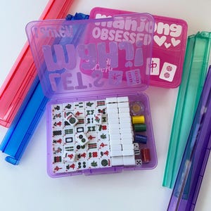May include: A purple Mahjong set with white tiles, dice, and colorful chips, inside a clear plastic case. The case has the text "Mahjong OBSESSED" and "WANT IT!" in a playful font. Additional game pieces are visible.
