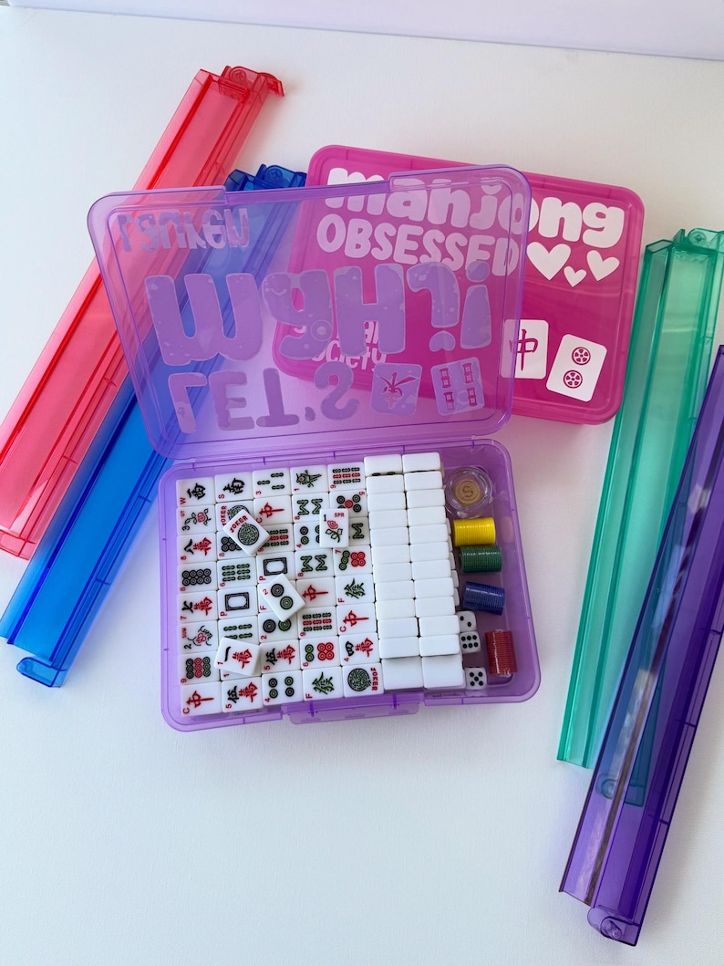 May include: A purple Mahjong set with white tiles, dice, and colorful chips, inside a clear plastic case. The case has the text "Mahjong OBSESSED" and "WANT IT!" in a playful font. Additional game pieces are visible.