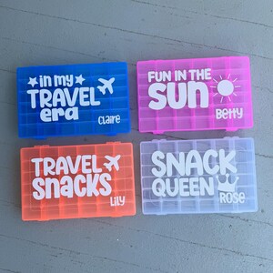 May include: Four brightly coloured plastic travel snack boxes. The boxes are blue, pink, orange, and white. Each box has white text. The blue box says "in my TRAVEL era Claire". The pink box says "FUN IN THE sun Betty". The orange box says "TRAVEL SNACKS Lily". The white box says "SNACK QUEEN Rose".