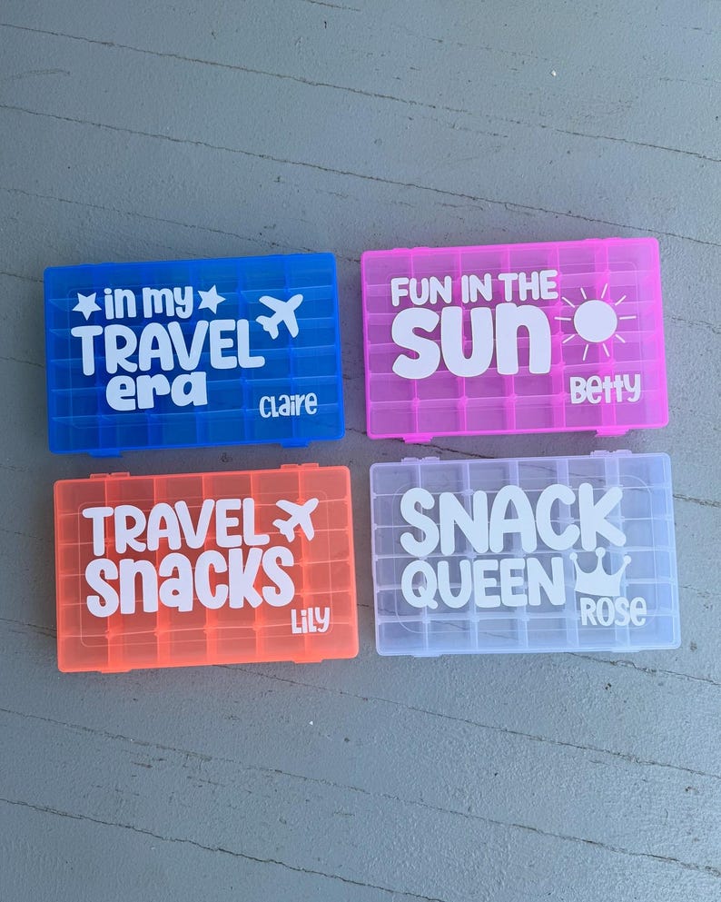 May include: Four brightly coloured plastic travel snack boxes. The boxes are blue, pink, orange, and white. Each box has white text. The blue box says "in my TRAVEL era Claire". The pink box says "FUN IN THE sun Betty". The orange box says "TRAVEL SNACKS Lily". The white box says "SNACK QUEEN Rose".