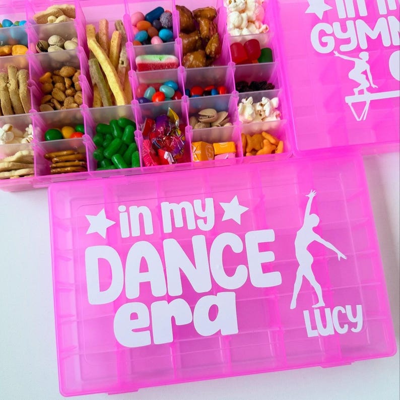 May include: A pink plastic snack organiser filled with a variety of sweets, nuts, and crackers. The organiser has multiple compartments and the text "in my DANCE era" in white. A dancer silhouette is also visible.