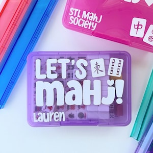 Puede incluir: Una colección de estuches de juego de Mahjong de colores. El estuche principal es morado con el texto "LET'S Mahj!" y "Lauren" en blanco. Otros estuches son rosas, azules y verdes. El estuche rosa tiene impreso "Mahjong OBSESSED" y "STL Mahj Society".