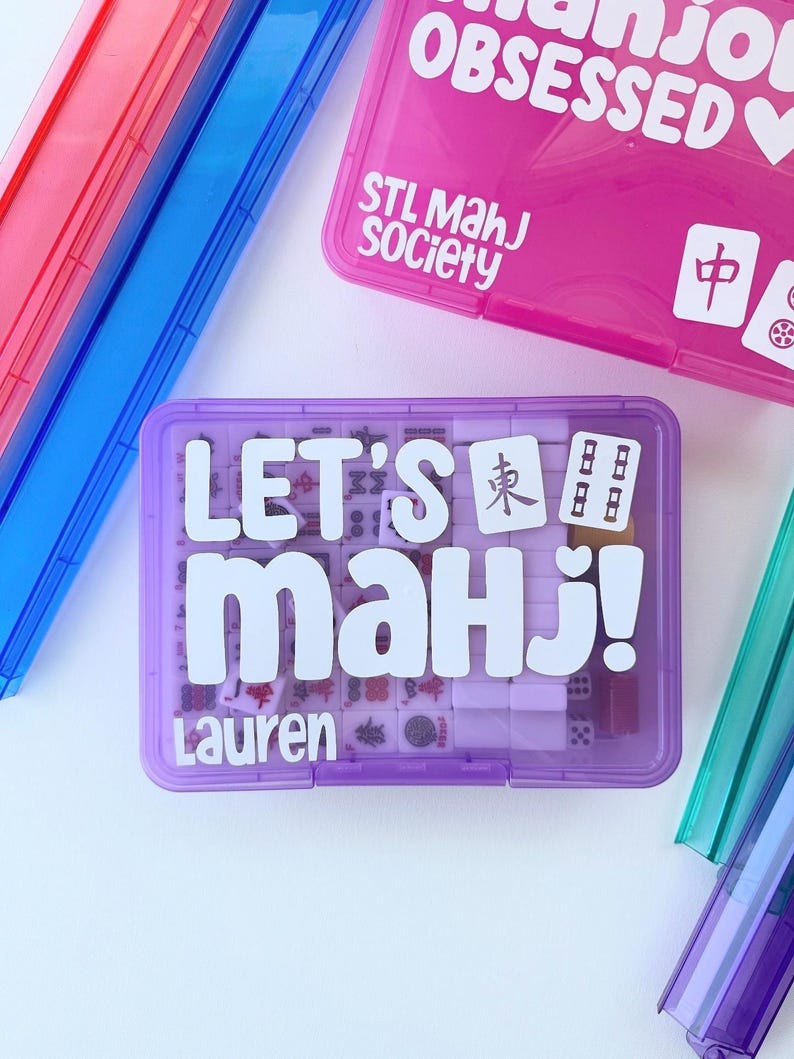 May include: A collection of colorful Mahjong game cases. The primary case is purple with the text "LET'S Mahj!" and "Lauren" in white. Other cases are pink, blue, and green. The pink case has "Mahjong OBSESSED" and "STL Mahj Society" printed on it.
