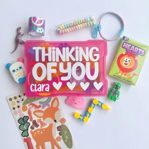 May include: A pink plastic storage box with the white text "THINKING OF YOU" and "Clara" with four hearts, surrounded by a sheet of deer stickers, a Hearts card game deck, a small dinosaur figure, sweets, a blue zipper bracelet, and toys.