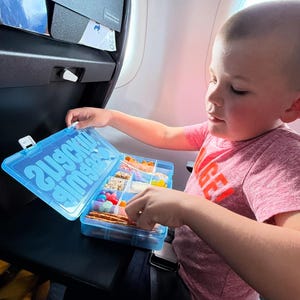 Peut inclure: Un récipient en plastique bleu avec les mots "Snack Time" et "Snacks" imprimés sur le couvercle. Le récipient est rempli de diverses collations, notamment des bretzels, des bonbons et des craquelins. Un enfant tend la main dans le récipient pour prendre une collation.
