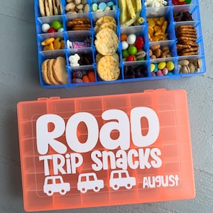 Peut inclure: Deux boîtes de collations en plastique remplies de diverses friandises. Le récipient supérieur est bleu et le fond est orange, avec les mots "ROAD TRIP SNACKS" et des graphiques de voiture. Le mot "August" est également visible.