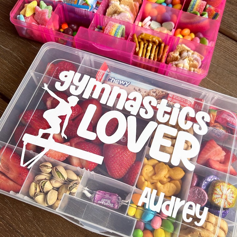 May include: Two clear plastic snack boxes with compartments. The bottom box has the words "gymnastics LOVER" and the name "Audrey" in white. It contains strawberries, pistachios, and snacks. The top box is pink and filled with colourful sweets and snacks.