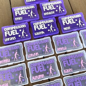 May include: Purple and clear plastic boxes with the words "Competition Fuel" and a gymnast silhouette. Each box has a different name in white or purple, such as "Evianna" and "Emily".