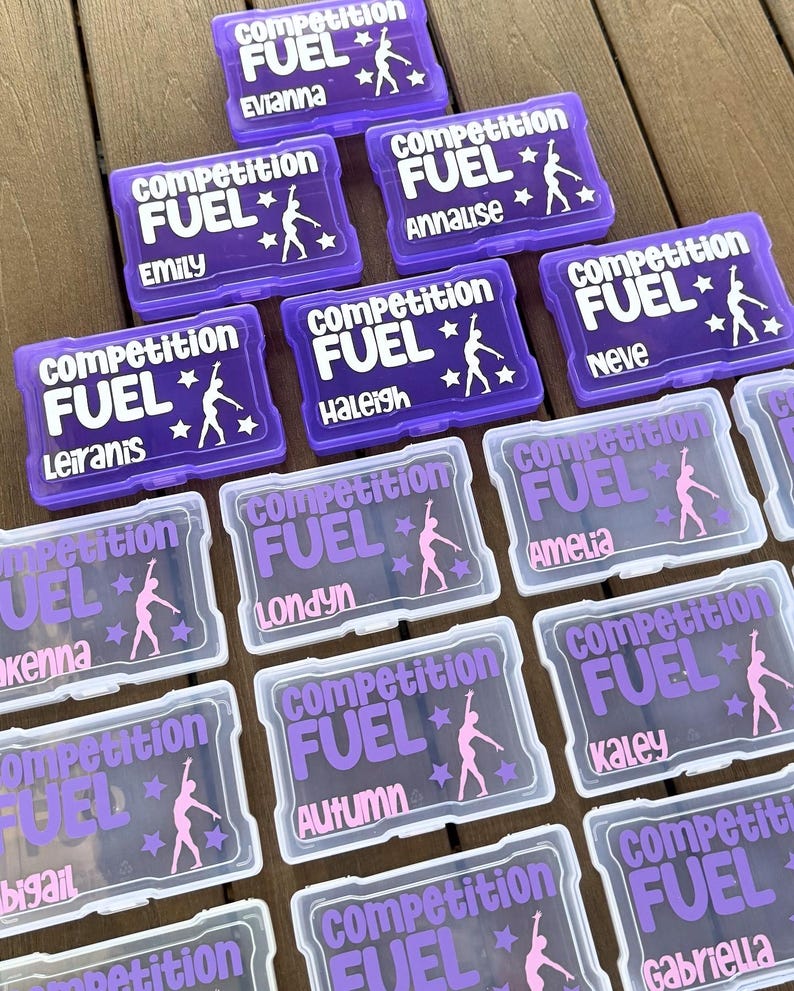 May include: Purple and clear plastic boxes with the words "Competition Fuel" and a gymnast silhouette. Each box has a different name in white or purple, such as "Evianna" and "Emily".