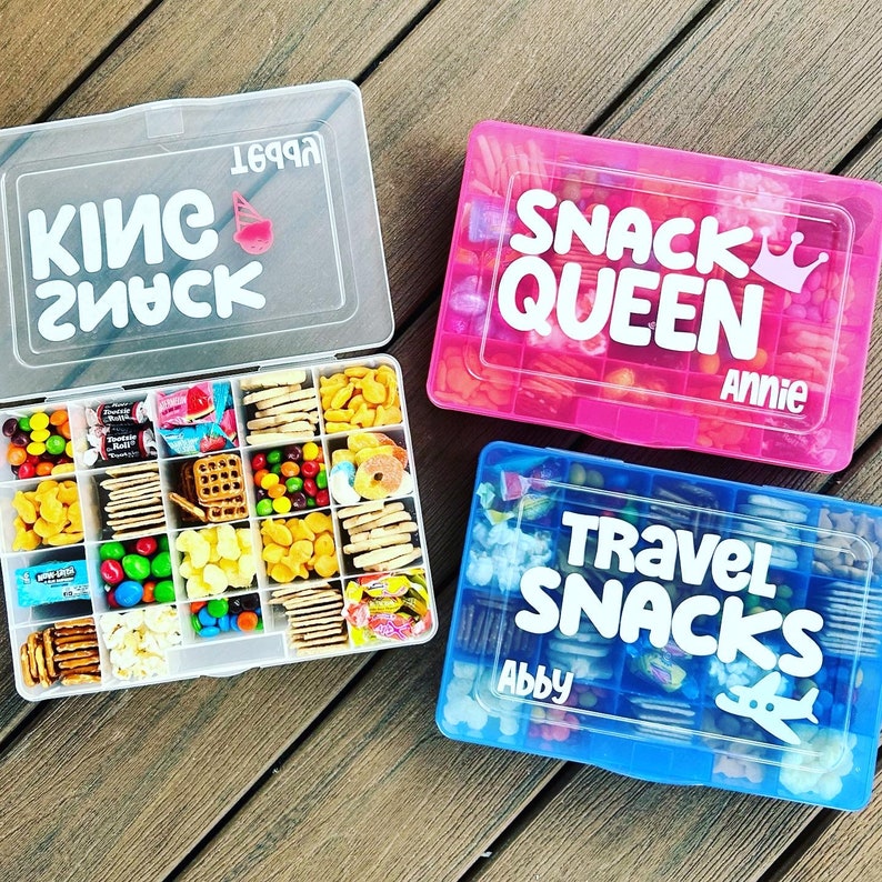 3 travel snack boxes. Clear box open with snacks inside each compartment. Goldfish crackers, skittles, tootsie rolls, pretzels, animal crackers, candy inside. Blue box with travel snack, name Abby and airplane. Pink Snack Queen box with crown.