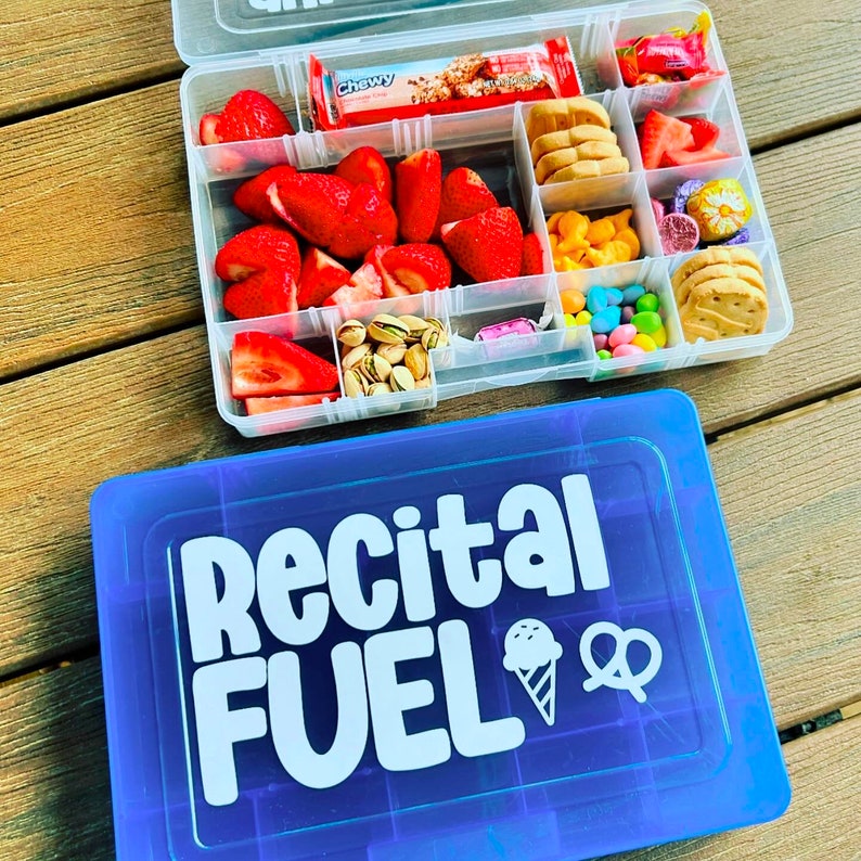 May include: A clear plastic container with compartments filled with snacks like strawberries, biscuits, sweets, and a granola bar. A blue container with "Recital FUEL" in white lettering is below.