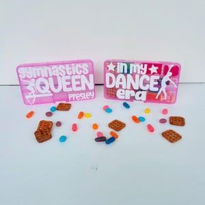 May include: Two pink plastic boxes with white text and small compartments. One says "gymnastics QUEEN Presley" and the other says "in my DANCE era". Small snacks and sweets are scattered around the boxes.