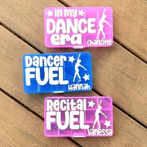 May include: Three colourful pill organisers with white text and dancer silhouettes. The pink organiser says "In my DANCE era" and "Charlotte". The blue organiser says "Dancer FUEL" and "Hannah". The pink organiser says "Recital FUEL" and "Vanessa".