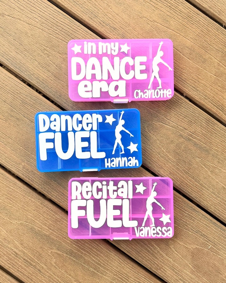 May include: Three colourful pill organisers with white text and dancer silhouettes. The pink organiser says "In my DANCE era" and "Charlotte". The blue organiser says "Dancer FUEL" and "Hannah". The pink organiser says "Recital FUEL" and "Vanessa".