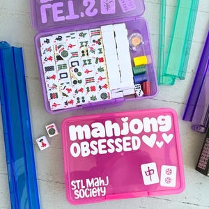 May include: A purple Mahjong set with white tiles, dice, and accessories. The lid has the text "W9H? TEL.2" in white. A pink case with "mahjong OBSESSED" and "STL MahJ Society" in white text is also visible.