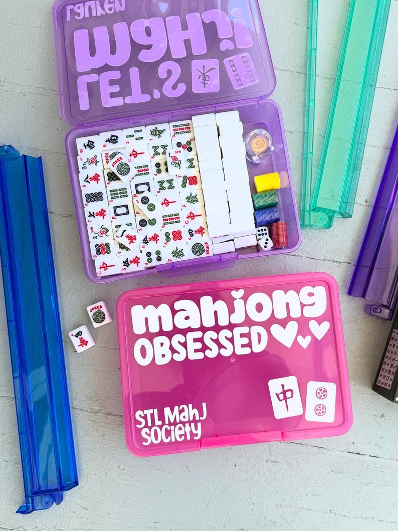 May include: A purple Mahjong set with white tiles, dice, and accessories. The lid has the text "W9H? TEL.2" in white. A pink case with "mahjong OBSESSED" and "STL MahJ Society" in white text is also visible.