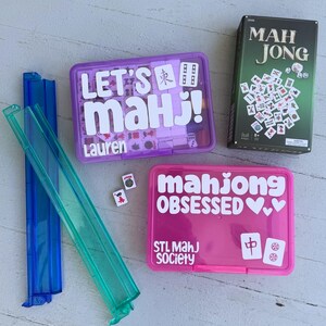May include: A collection of Mahjong game sets and accessories. Includes a green box labeled "MAH JONG", two plastic tile racks, and two plastic cases with the text "LET'S MAH J!" and "mahjong OBSESSED".