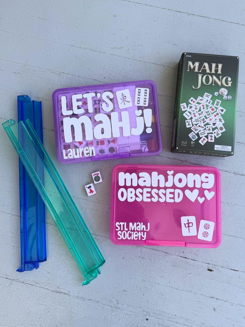 May include: A collection of Mahjong game sets and accessories. Includes a green box labeled "MAH JONG", two plastic tile racks, and two plastic cases with the text "LET'S MAH J!" and "mahjong OBSESSED".