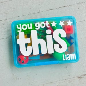 Puede incluir: Un contenedor de almacenamiento rectangular de plástico azul translúcido lleno de varios juguetes sensoriales coloridos. La tapa presenta texto blanco que dice "you got this" con tres estrellas y el nombre "Liam" en la esquina inferior derecha.