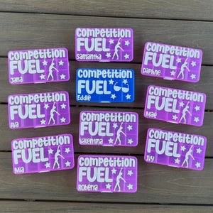 Pode incluir: Uma cole&ccedil;&atilde;o de nove recipientes retangulares de pl&aacute;stico para lanches em roxo e azul. Cada recipiente apresenta o texto branco "competition FUEL" com uma silhueta de ginasta e estrelas. Nomes individuais est&atilde;o impressos em cada um, incluindo Jana, Samantha, Daphne, Ava, Eddie, Mae, Valentina, Mia e Roberta.